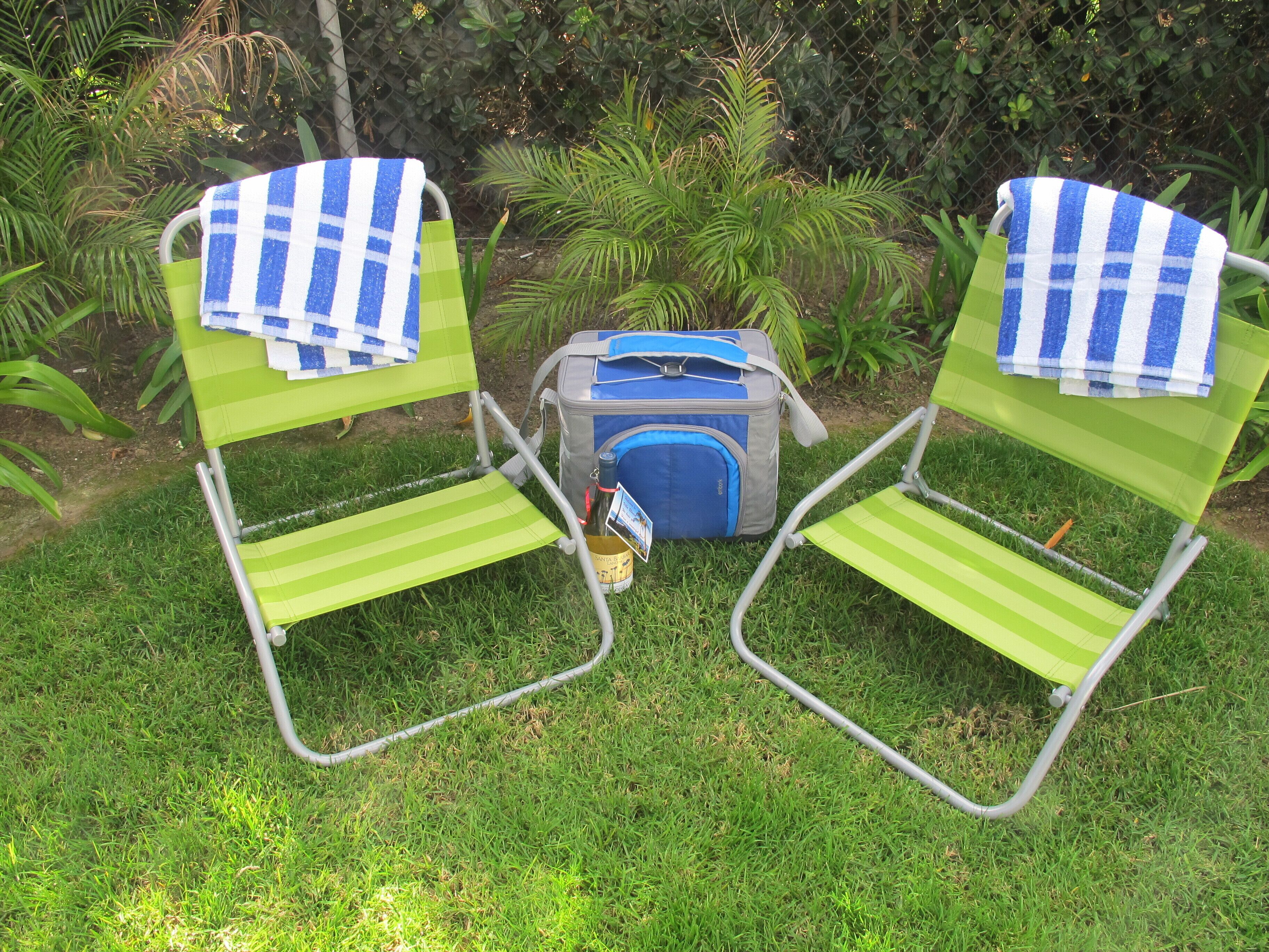 Two green lawn chairs with blue and white checkered towels, a gray backpack, and a bottle of water on a grassy area with plants in the background.