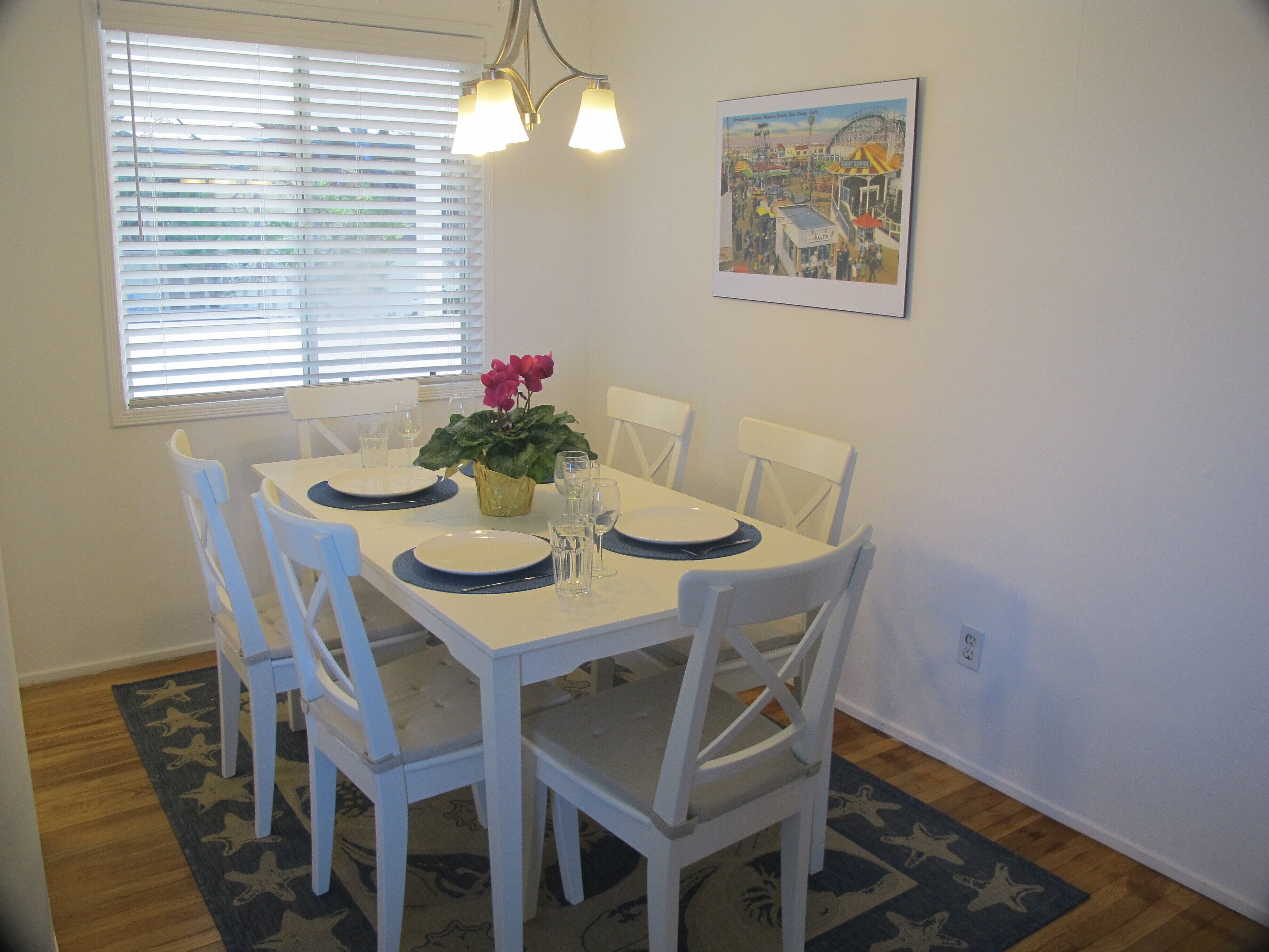 A dining area with a white table set for four, surrounded by white chairs. There is a window with blinds, a painting on the wall, and a plant on the table.