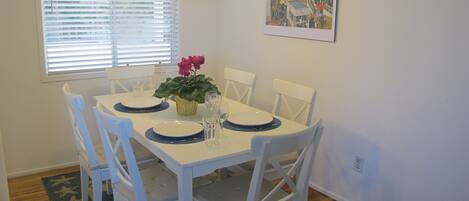 A dining area with a white table set for four, surrounded by white chairs. There is a window with blinds, a painting on the wall, and a plant on the table.