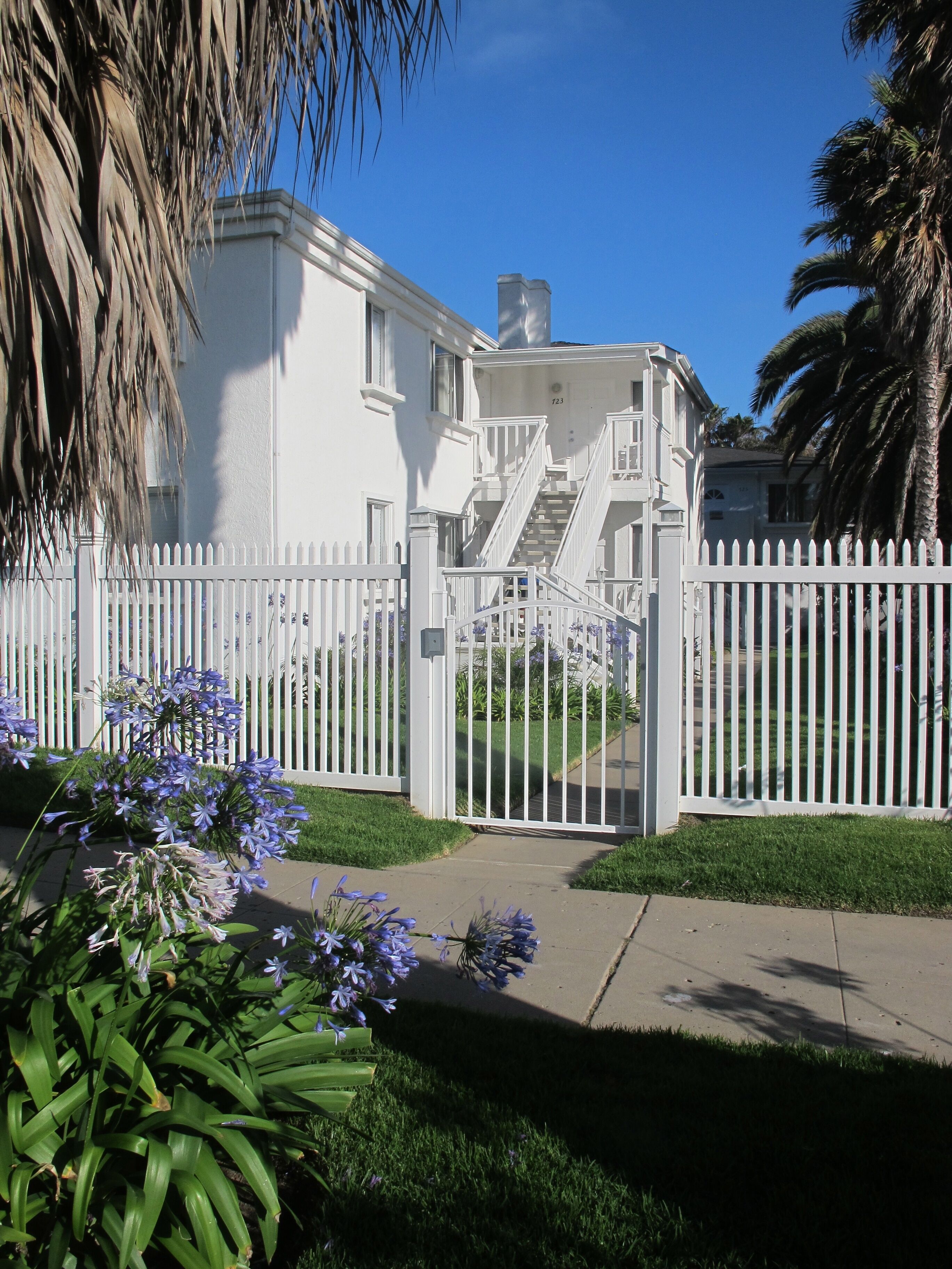 Une maison blanche à deux étages, avec une clôture en bois peinte en blanc, un escalier extérieur bien visible et une pelouse bien entretenue.