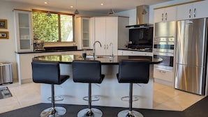 A modern kitchen with a central island, stainless steel appliances, and black bar stools. - BELVEDERE-(meaning in Italian PANORAMIC VIEWS) 3 BEDROOM HOUSE IN QUEENSTOWN (Fernhill)