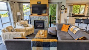 A living room with a stone fireplace, a grey sofa, a beige armchair, and a coffee table. - BELVEDERE-(meaning in Italian PANORAMIC VIEWS) 3 BEDROOM HOUSE IN QUEENSTOWN (Fernhill)