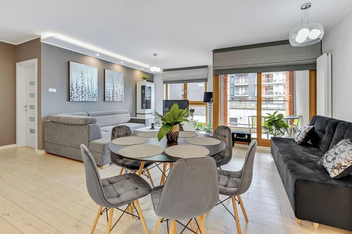 Una sala de estar moderna con un sofá gris, un sofá negro, una mesa de comedor con sillas y un ventanal con vistas a edificios.