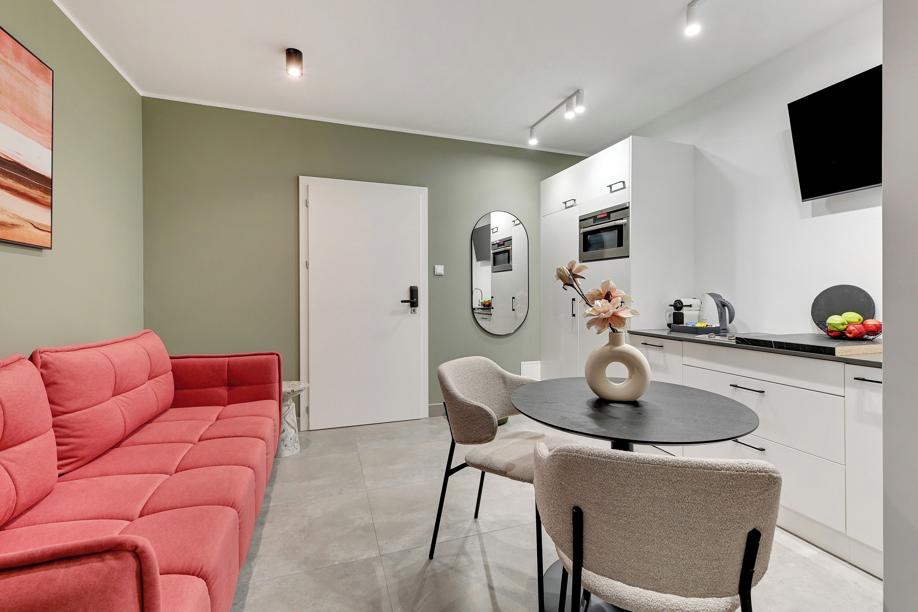 A modern living room with a red sofa, beige chairs, and a round table with a vase. A kitchen with white cabinets and a TV is visible in the background.