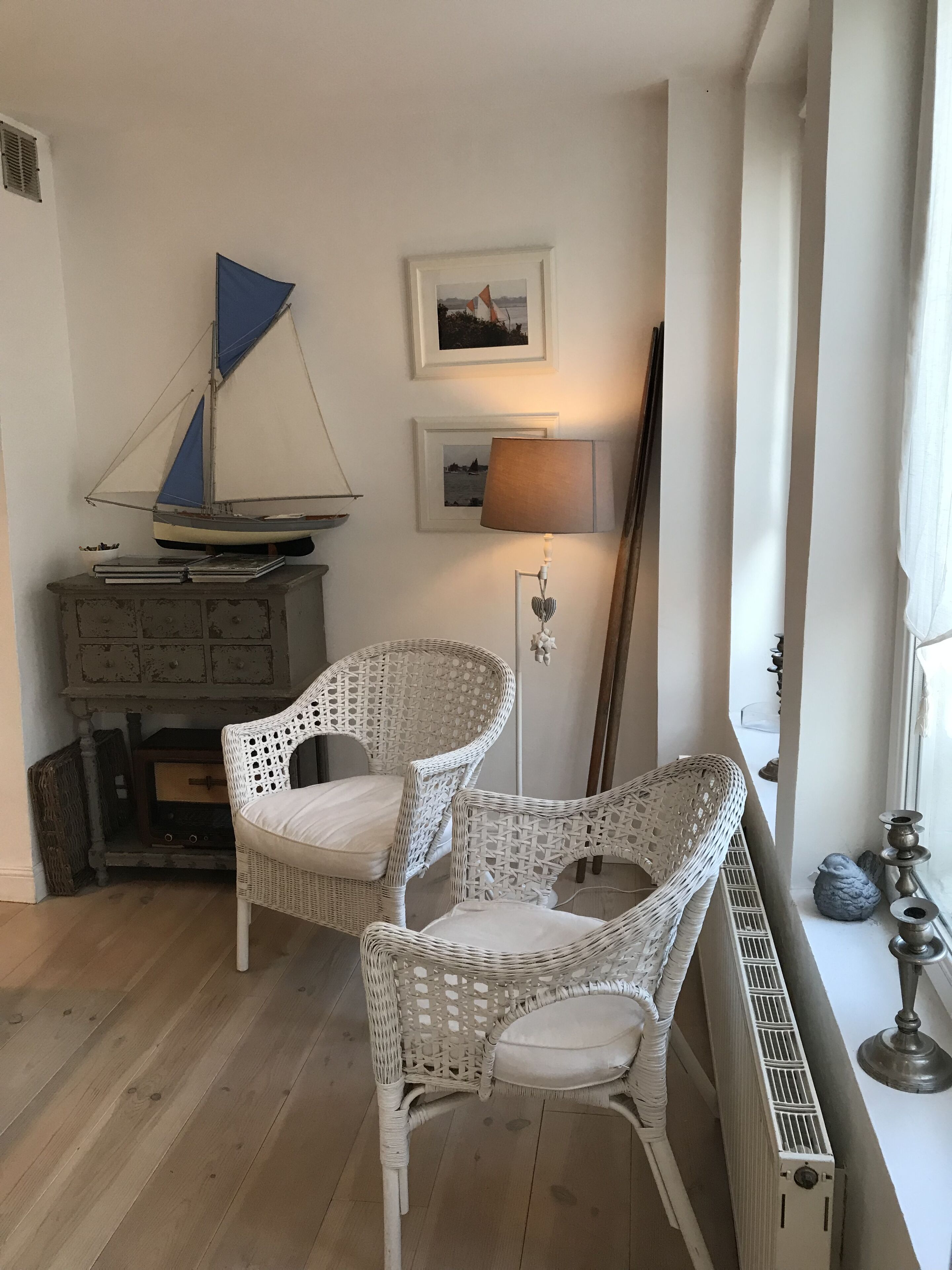 Quarto com duas cadeiras de vime, um pequeno modelo de barco à vela, uma luminária e quadros nas paredes.