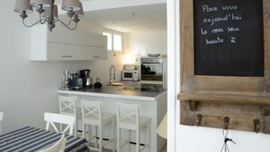 Una cocina moderna con armarios blancos, una isla central con taburetes y una lámpara colgante. Un pizarrón con escritura francesa está fijado en la pared.