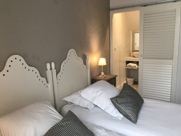 A bedroom with a bed, pillows, a nightstand with a lamp, and a bathroom visible through an open door.