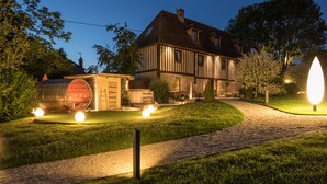 Une grande maison à deux étages, dotée d’un toit en pente, d’une cheminée bien visible et d’une pelouse bien entretenue. Un chemin pavé mène à la maison, et sur la gauche, on aperçoit un grand objet éclairé qui ressemble à une cuve ou à un silo.