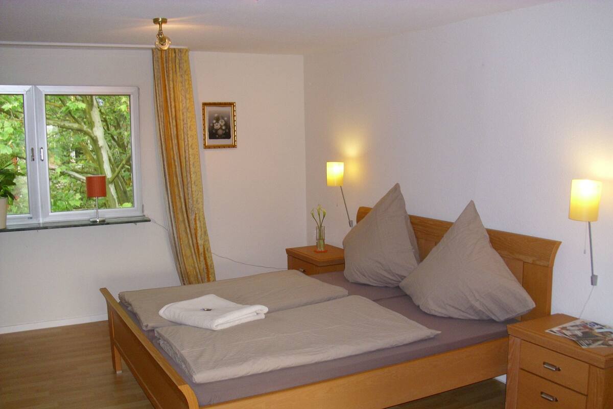 En snyggt bäddad säng med kuddar och en hopvikt handduk, ett sängbord med en lampa, ett litet bord bredvid sängen med en tidning och ett fönster med en gardin.