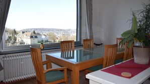A dining area with a glass table and wooden chairs, a potted plant, and a view of a cityscape through a large window. - Messewohnung Augusta (Wuppertal)