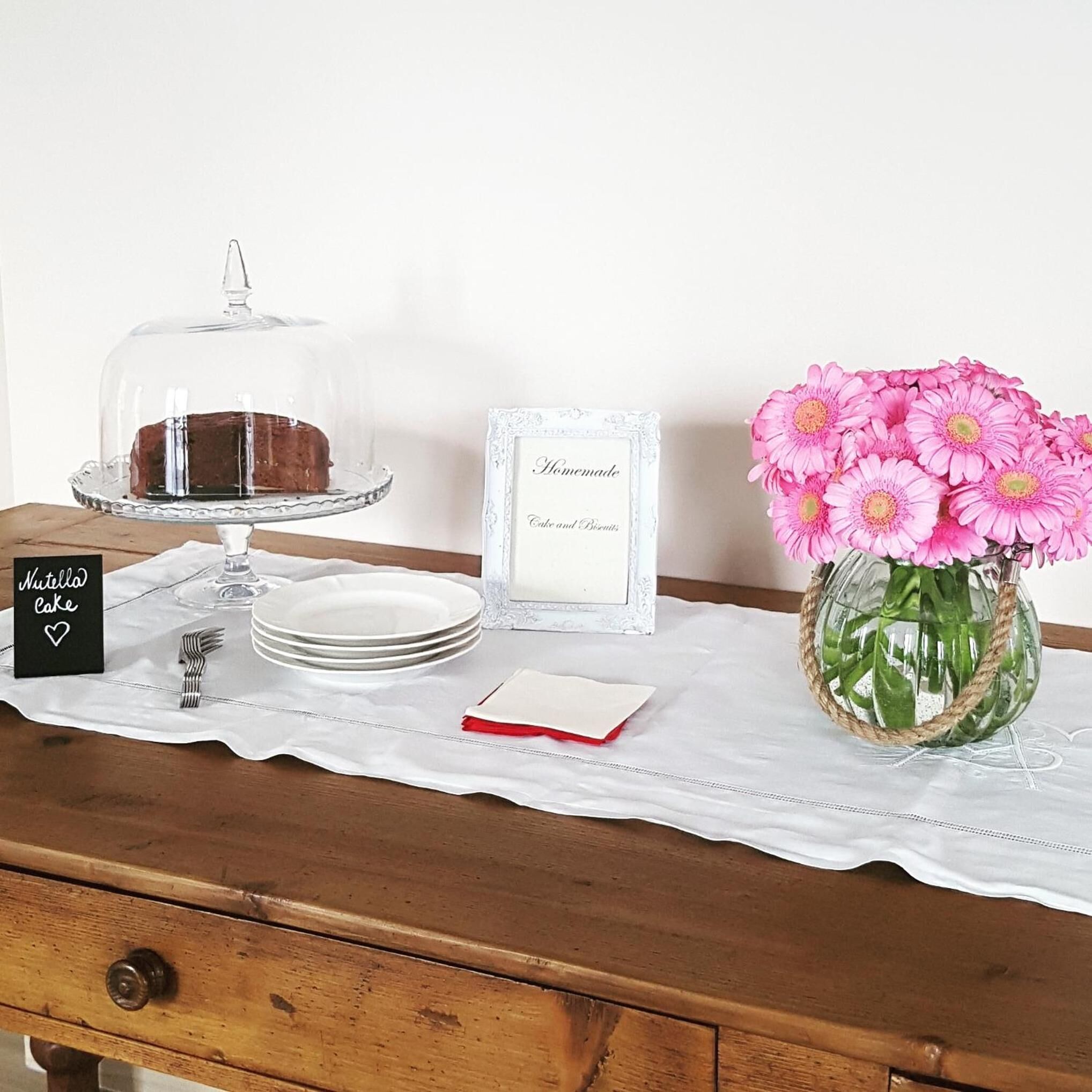 A dessert table with a cake under a glass dome, a sign labeled "Nutella Cake," plates, a napkin, and a framed sign reading "Homemade Cake and Biscuits."