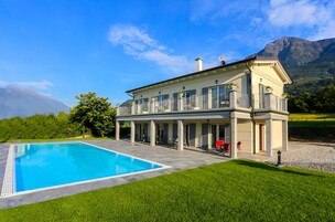 Uma casa de dois andares com piscina, varanda coberta e montanhas ao fundo.