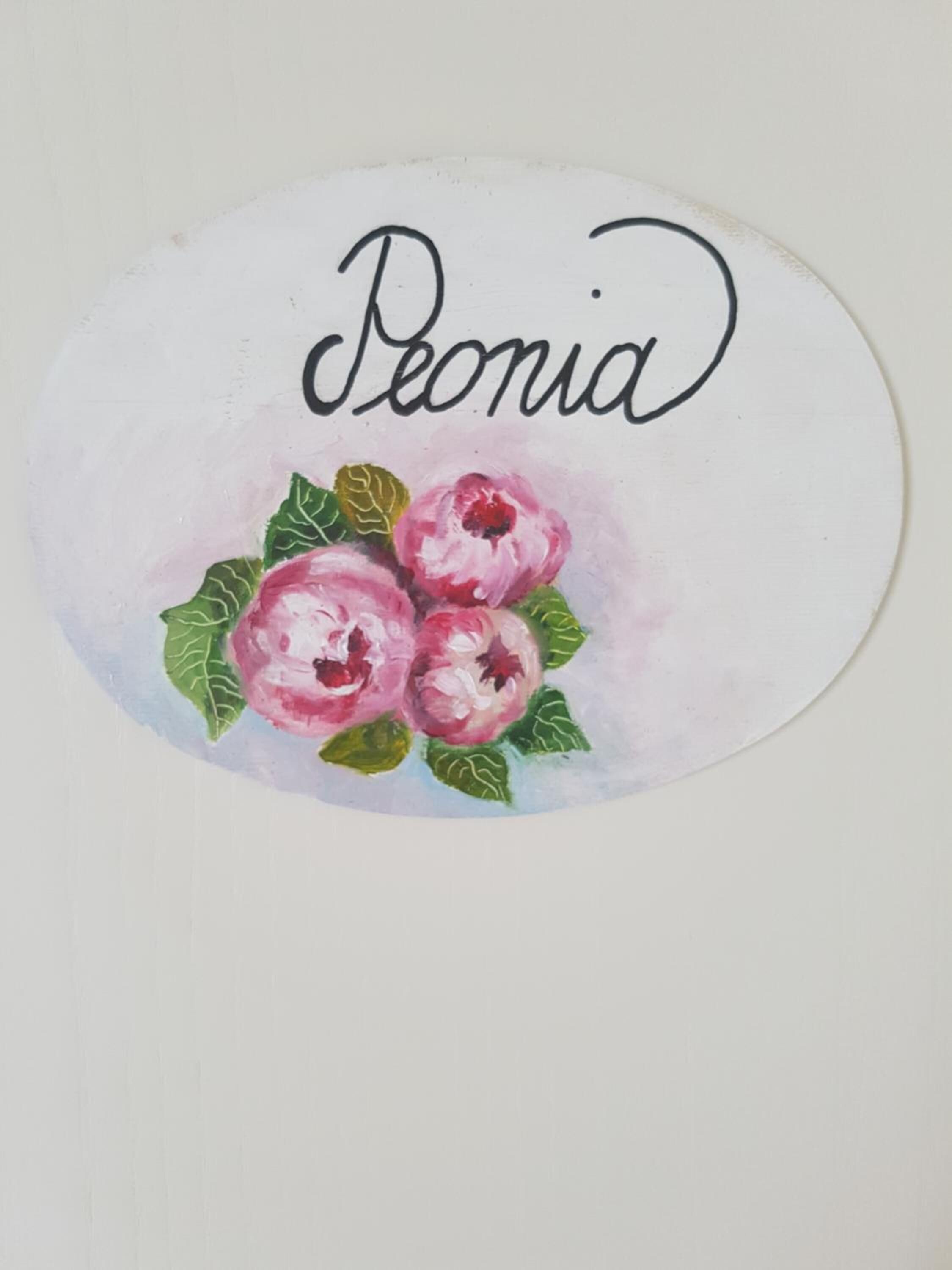 Un panneau circulaire portant le nom « Peonia » et une aquarelle représentant trois fleurs roses aux feuilles vertes.