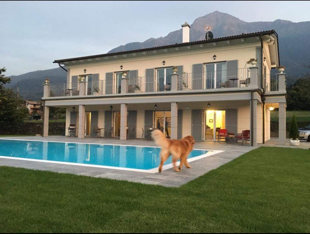 Una casa grande de dos pisos con piscina, un césped cuidado y montañas al fondo.