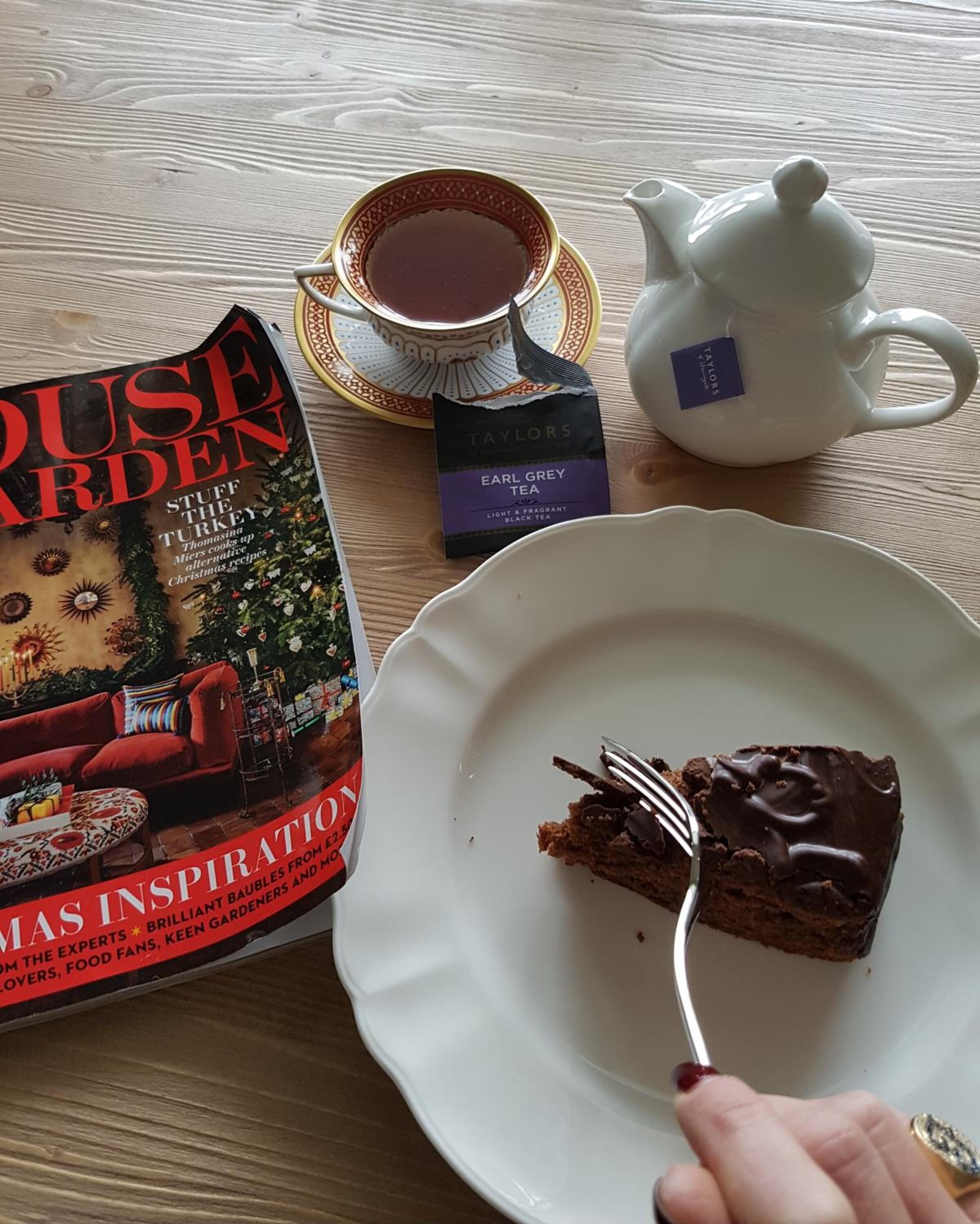 A table with a book titled 'House Garden', a teapot, a cup of tea, and a slice of chocolate cake on a plate.