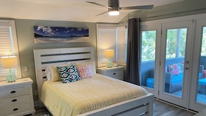 A bedroom with a bed, nightstands, a ceiling fan, a window with blinds, and a view of an outdoor seating area. - Sea Salt Cottage - Beach front neighborhood, next to downtown restaurants (Isle of Palms)