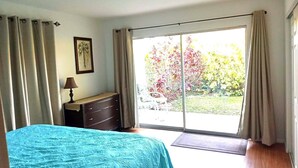 Un dormitorio con cama, cómoda, lámpara y vista a un jardín por una puerta corrediza de vidrio.