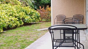 Un patio con mesa y sillas, rodeado de exuberante follaje y una pared color beige.