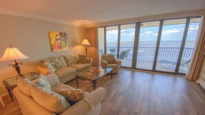 Ein geräumiges Wohnzimmer mit einem großen Fenster und Meerblick. Zwei Sofas, ein Couchtisch und eine Lampe sind vorhanden.