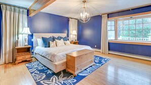 Quarto com cama grande, cabeceira de madeira, luminária pendente, mesa de cabeceira, cômoda e janela com vista para árvores.