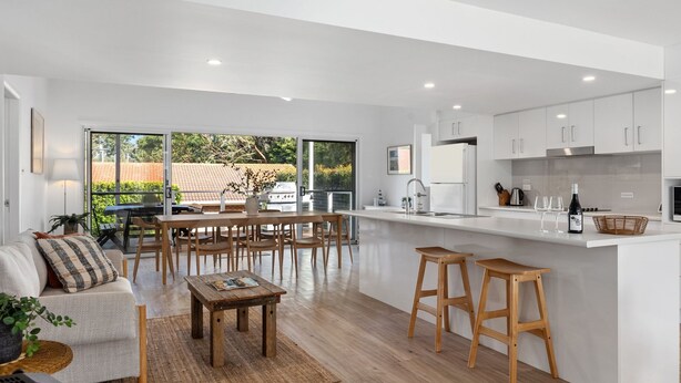 A modern kitchen with white cabinets, a central island, and a dining area with wooden furniture. - The Haven in Mollymook (Mollymook Beach)