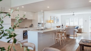 A modern kitchen with white cabinets, a central island with stools, and a dining area with a wooden table. - The Haven in Mollymook (Mollymook Beach)