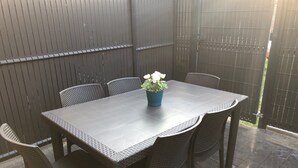 Un espacio al aire libre con una mesa oscura y sillas de mimbre, una planta en maceta con flores blancas y una reja metálica.
