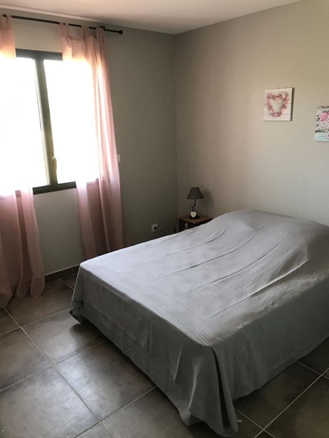 A bedroom with a bed, a nightstand, a lamp, a window with curtains, and a wall decoration.