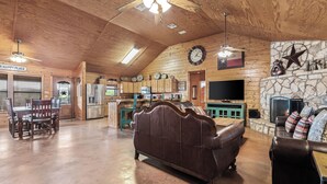 Un salón amplio con techo de madera, chimenea de piedra, televisor y zona de comedor con mobiliario de madera.