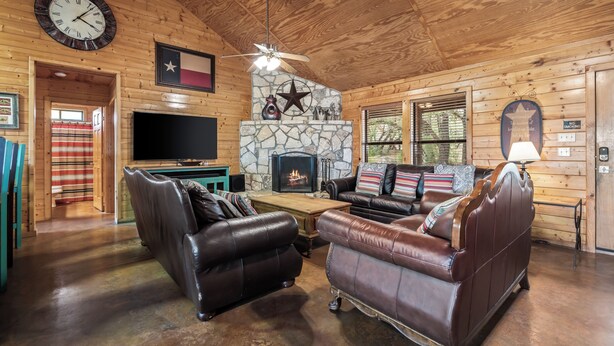 Un interior de cabaña acogedor con sofás de cuero, una chimenea de piedra, un televisor y una bandera de Texas.