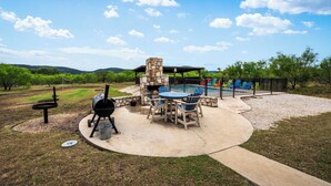 Un patio exterior con una parrilla de piedra, una mesa redonda con sillas y vistas a una piscina y colinas al fondo.
