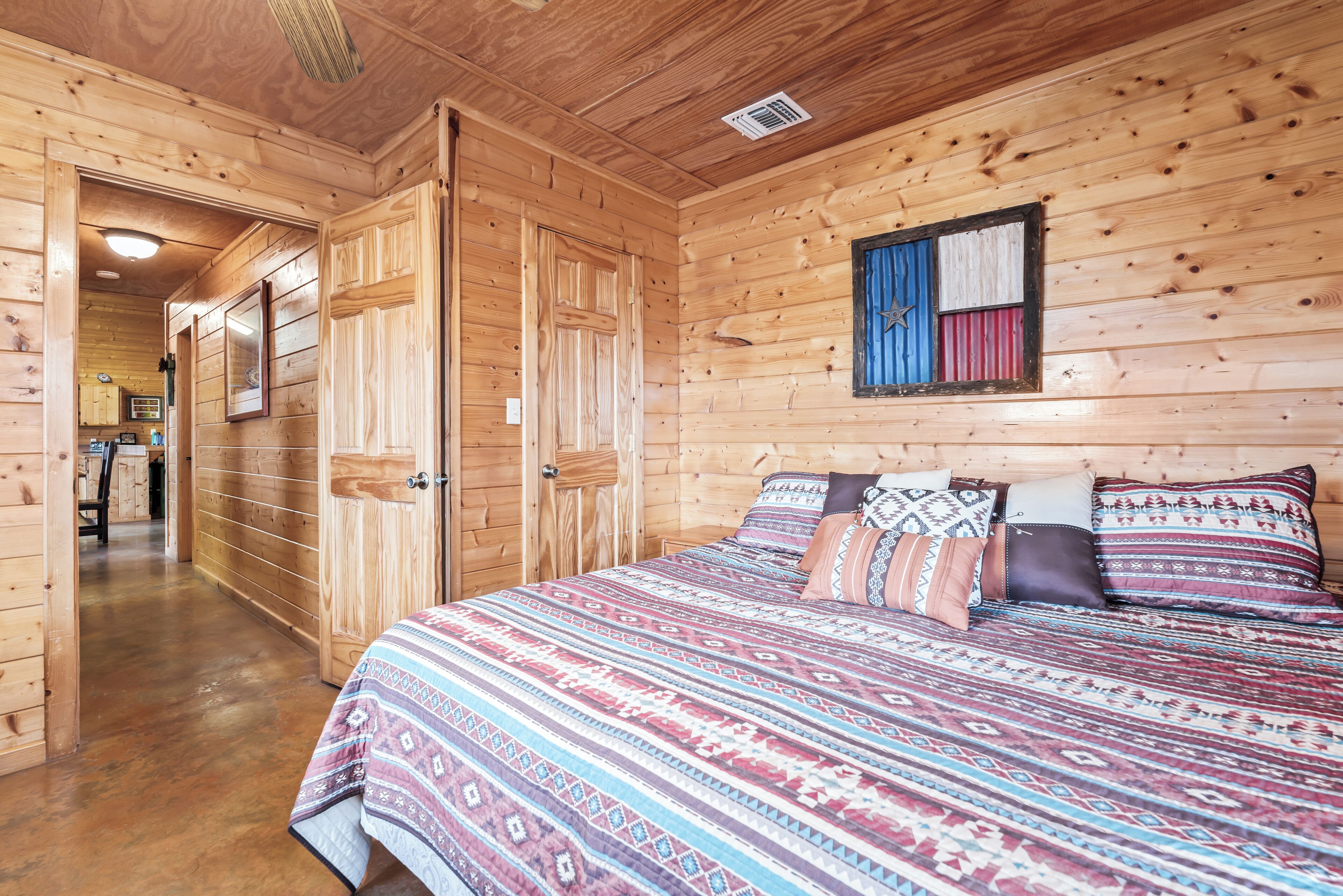 A wooden cabin interior with a large bed, wooden walls, and a hallway leading to a kitchen area.