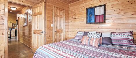 A wooden cabin interior with a large bed, wooden walls, and a hallway leading to a kitchen area.