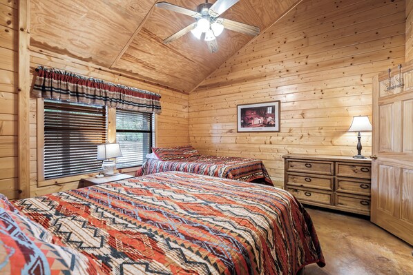 A wooden cabin bedroom with two beds, a ceiling fan, a window with blinds, a bedside table with a lamp, and a dresser. - Private pool, single story, NO bunk beds, less than 5-minute drive to THE Frio River in Concan, TX (Concan)