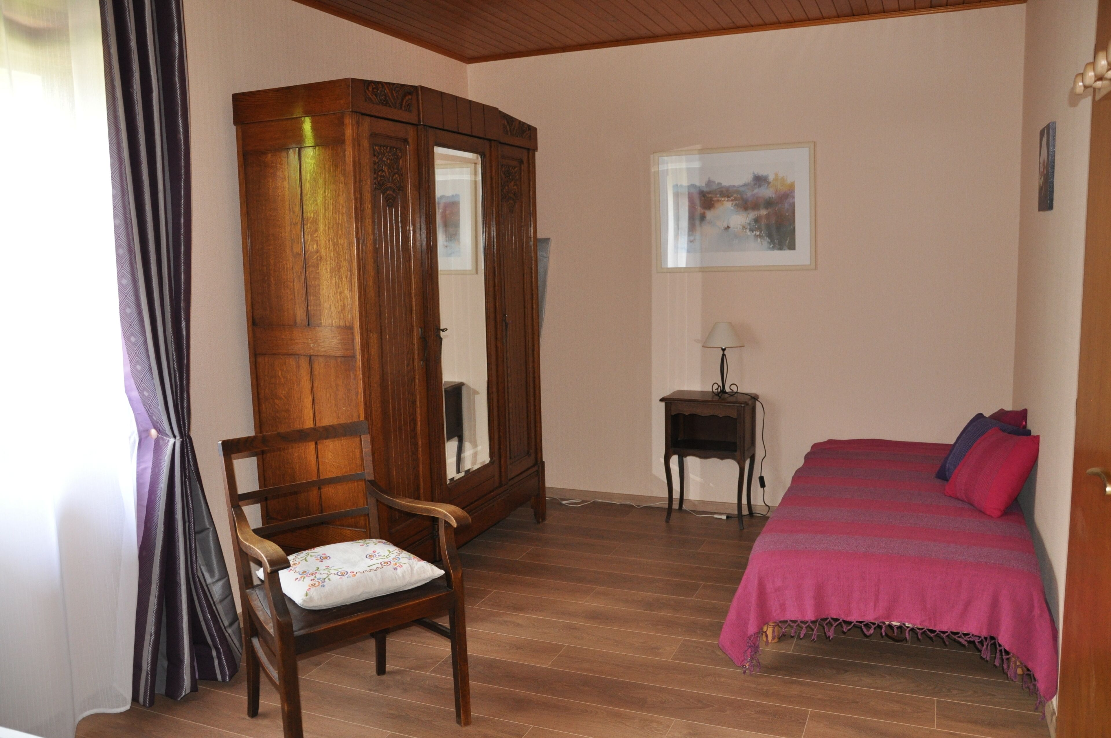 A bedroom with a wooden wardrobe, a bed with a red blanket, a chair with a cushion, a small table with a lamp, and a window with a painting.