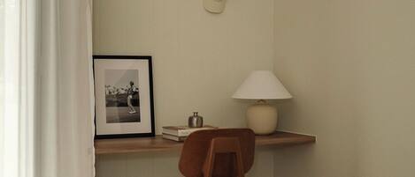 A hotel room with a bed, a chair, a desk with a lamp, a framed photo, and a wall-mounted hat.