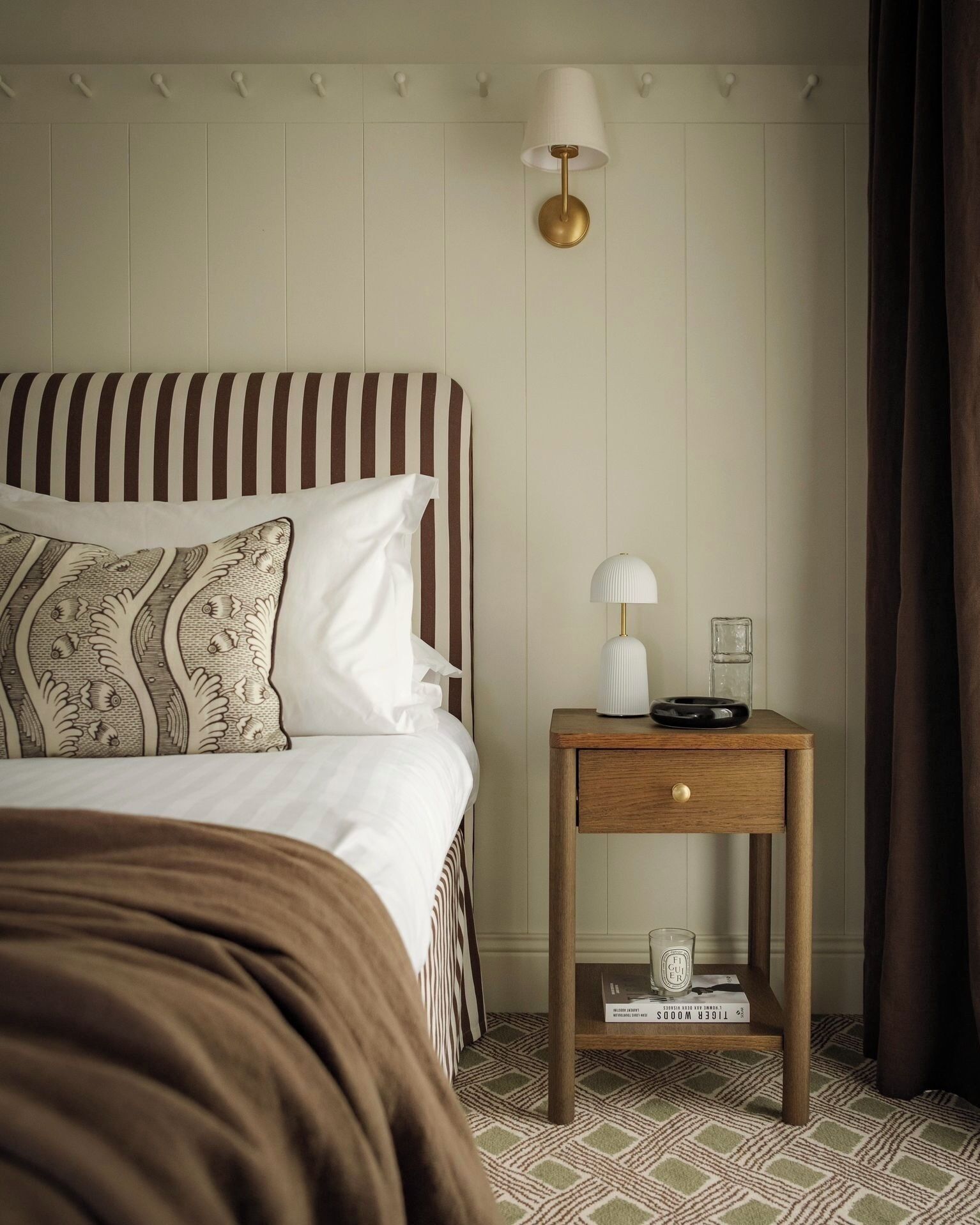 Una habitación de hotel con una cama, una mesita de noche con lámpara y una pequeña mesa con un vaso y un libro.