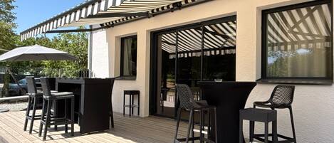 An outdoor patio with black bar stools and a large umbrella, a wooden deck, and a building with a striped awning.