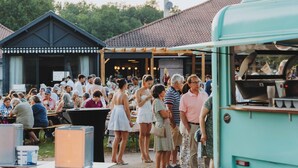 Un evento al aire libre concurrido, con gente de pie y sentada en mesas, un edificio azul y un autobús verde.