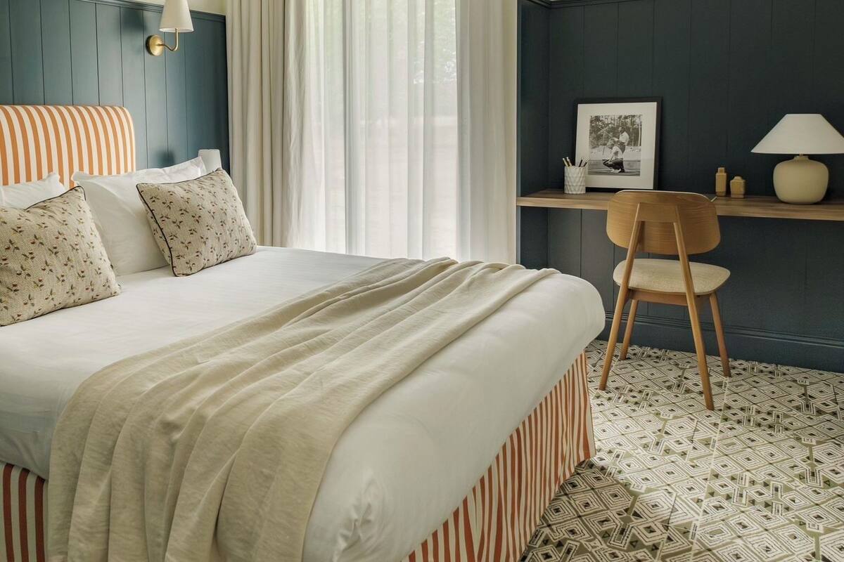 A hotel room with a bed, a desk, a chair, a window with curtains, and a wall-mounted lamp.