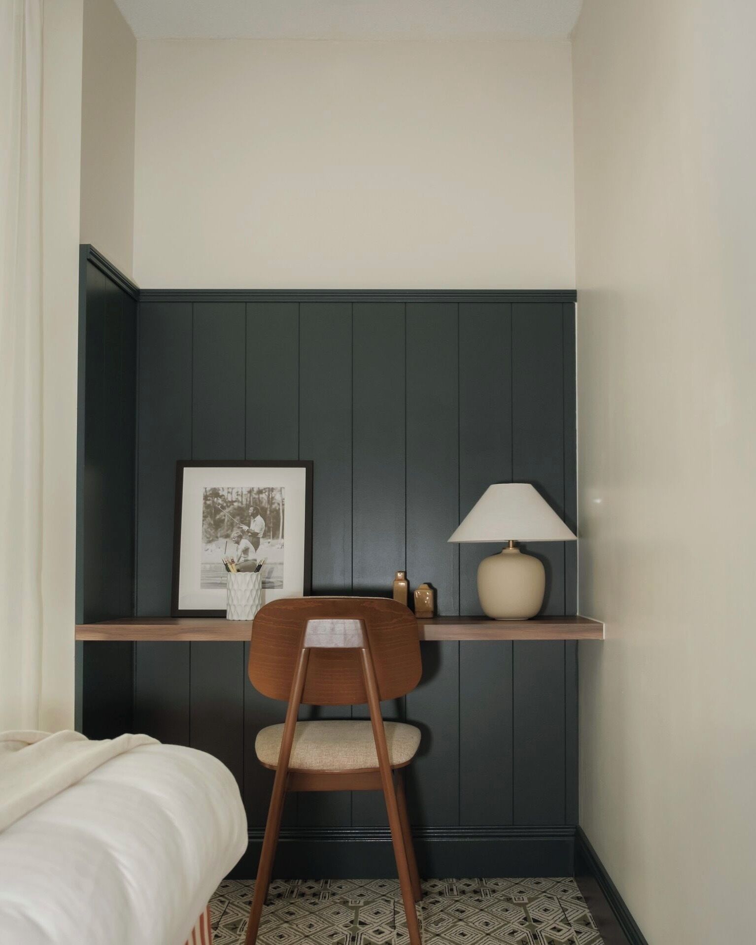 Een kleine, verzorgde kamer met een bed, een stoel, een bureau met een lamp en een lijstje aan de muur.