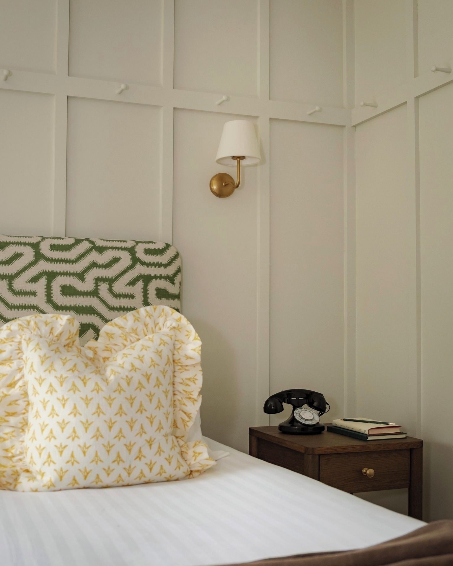 A neatly made bed with patterned pillows, a bedside lamp, a telephone, and a small wooden nightstand.