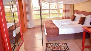 A hotel room with a large bed, a bedside table, a mirror, and a view of the outdoors. - Greenvale Hotel (Ruiru)