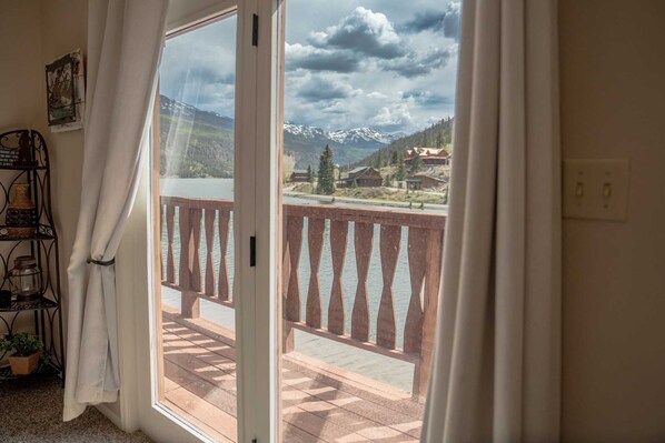 A room with a large sliding glass door leading to a balcony with a view of mountains and a lake.