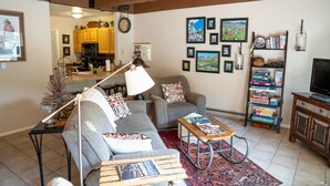Ein gemütliches Wohnzimmer mit einer Couch, einem Couchtisch und einem Bücherregal. Im Hintergrund ist eine Küche zu sehen.