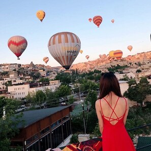 Une femme vêtue d’une robe rouge, qui contemple un paysage urbain avec de nombreux ballons à air chaud dans le ciel.