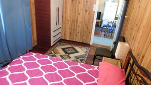 A bedroom with a bed, a wooden wardrobe, a small table with a chair, and a view into a kitchen area.
