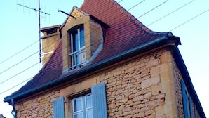 Un edificio de piedra con dos ventanas y contraventanas azules, que presenta un pequeño balcón y una chimenea.