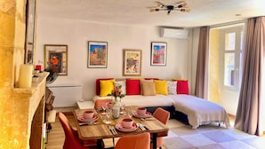 Una habitación con una mesa de comedor preparada para dos personas, un sofá con cojines coloridos, un ventilador de techo y cuadros en las paredes.