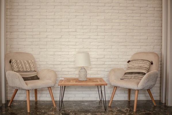 Two armchairs with patterned cushions, a small wooden table with a lamp, and a white brick wall. - Oikia Guesthouse (Kalabaka)